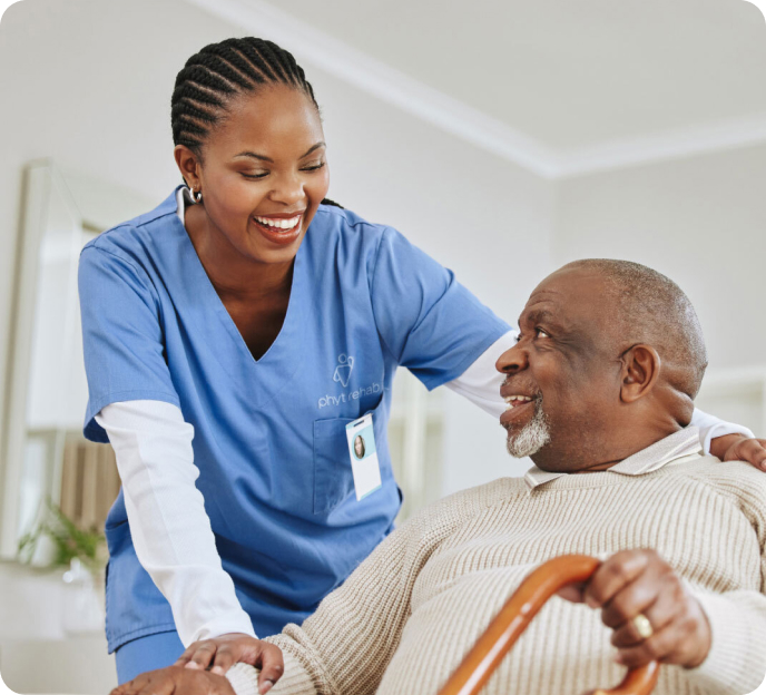 A healthcare worker in blue scrubs and a name badge is smiling and assisting an elderly man sitting with a cane.