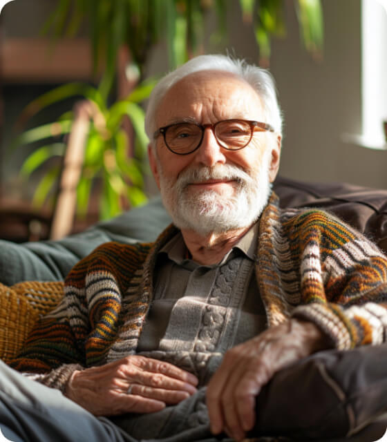 An elderly man with glasses and a sweater sitting comfortably on a couch.