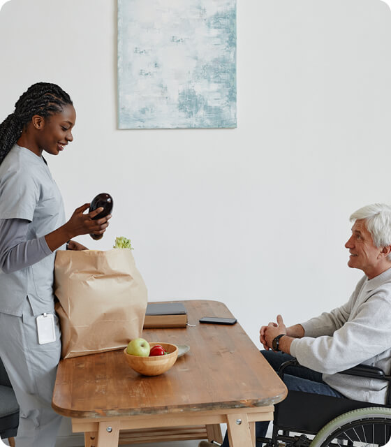 A healthcare worker unpacks groceries at a table while a man in a wheelchair looks on.