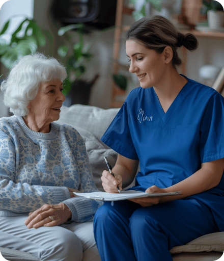 A woman in scrubs sits on a couch beside an older woman who is reading a book, creating a warm and caring atmosphere.